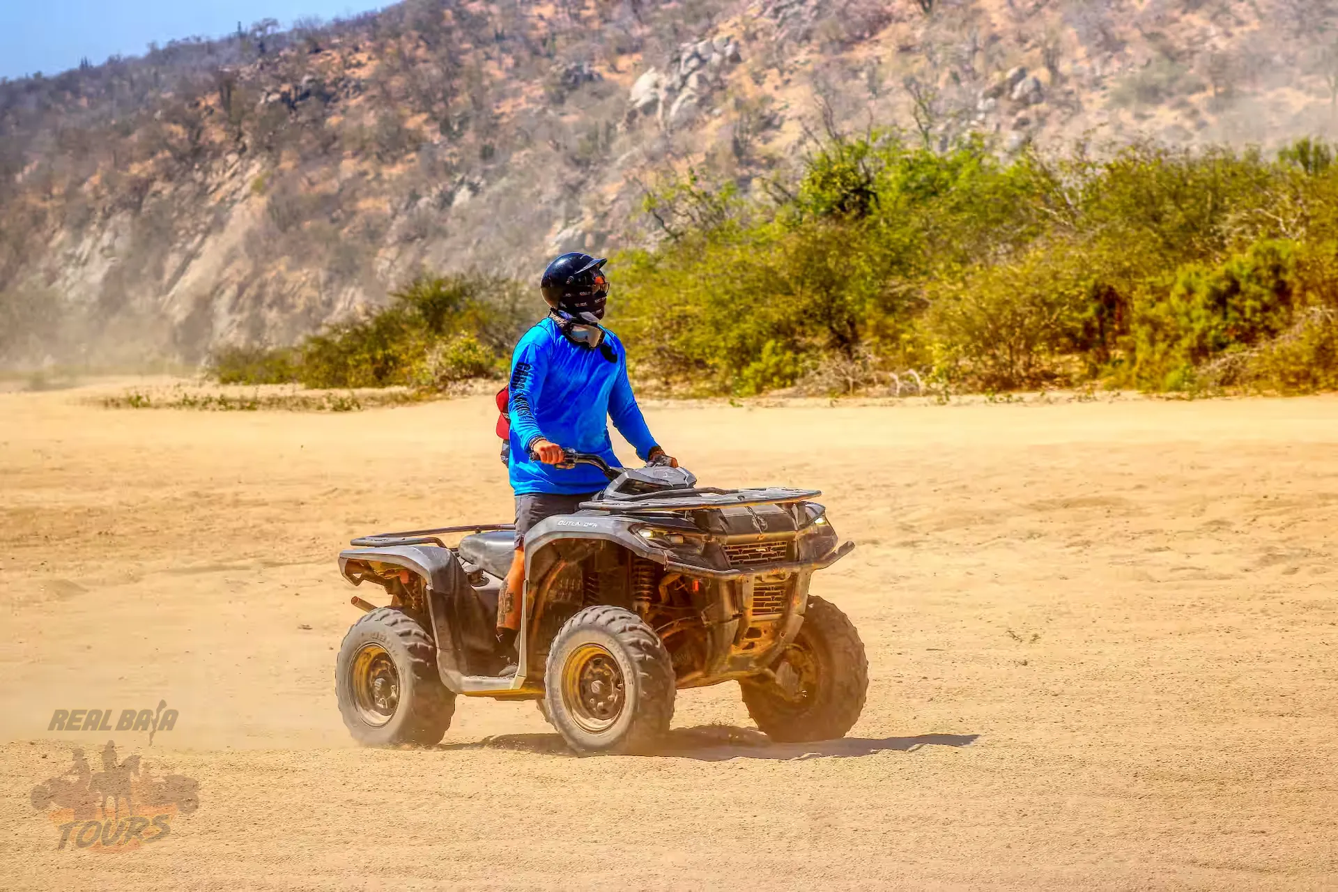 ATV piloto in blue jacket on dusty sendero del desierto with rocky hillside and green vegetation in Baja California Sur