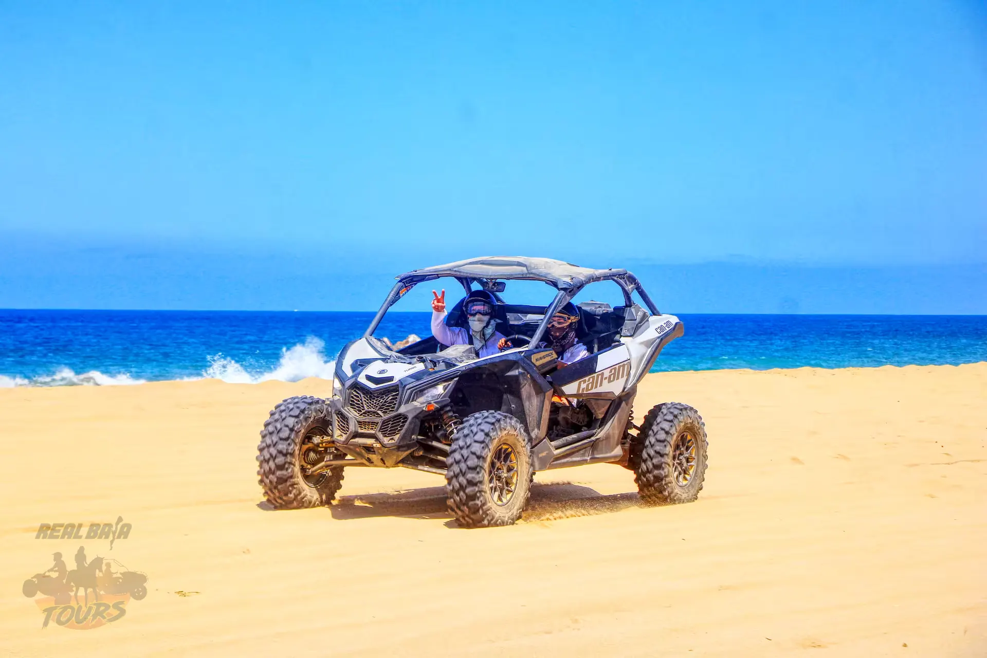 Two tourists driving a Can-Am Maverick side-by-side buggy on a playa de arena with turquoise olas del océano and blue sky in Baja California Sur Two tourists driving a Can-Am Maverick side-by-side buggy on a playa de arena with turquoise olas del océano and blue sky in Baja California Sur