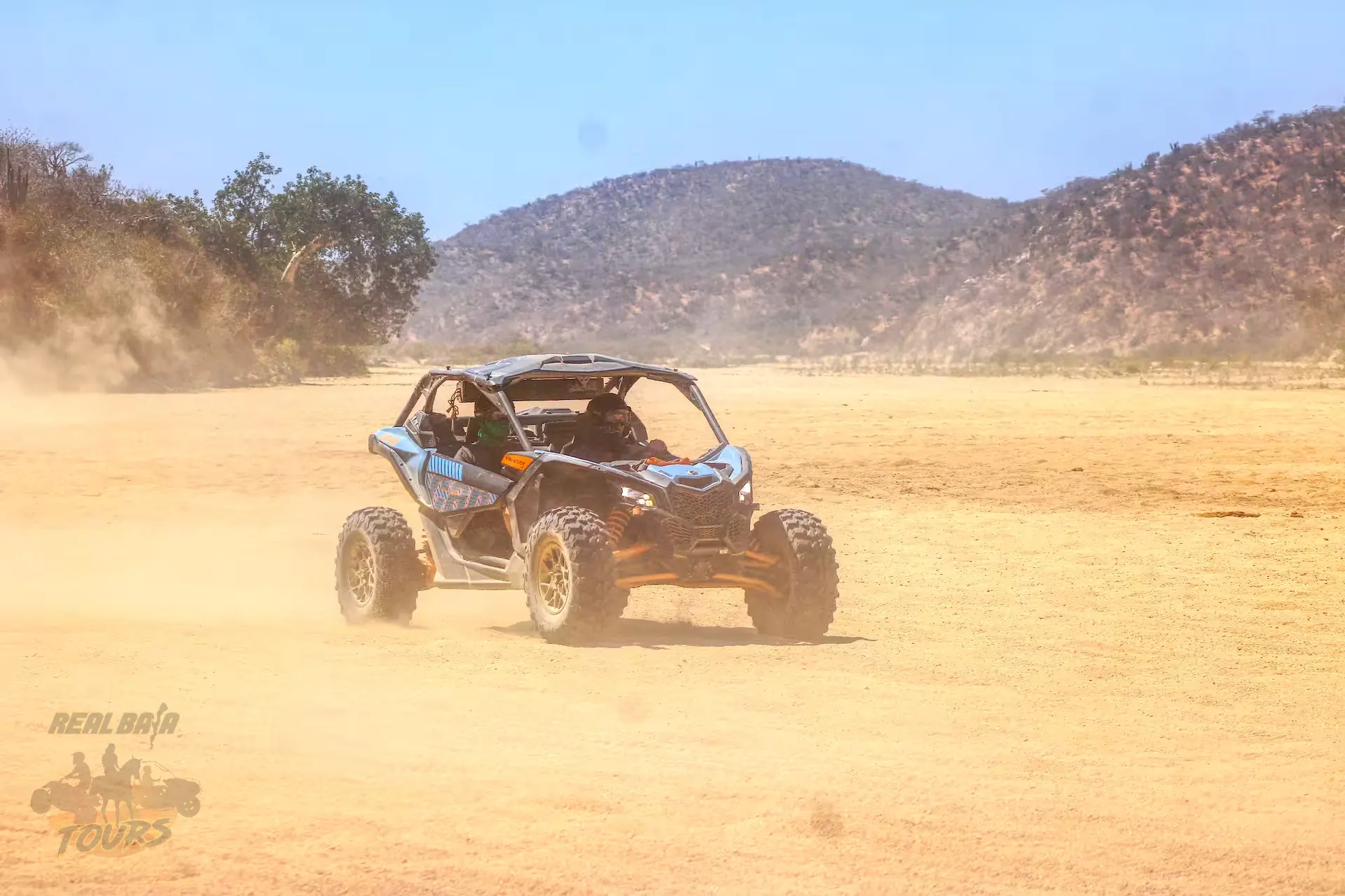 Blue and black UTV kicking up dust while driving across a sandy desert valley with mountains in Baja California Sur