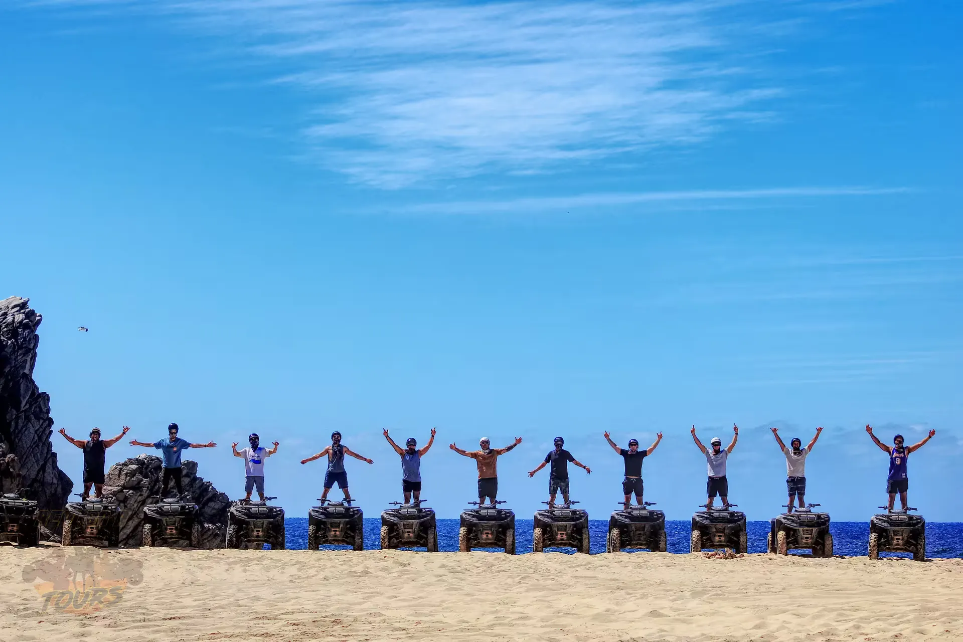 Group of tourists standing with arms raised on rocks at a Baja California Sur playa with blue ocean and sky