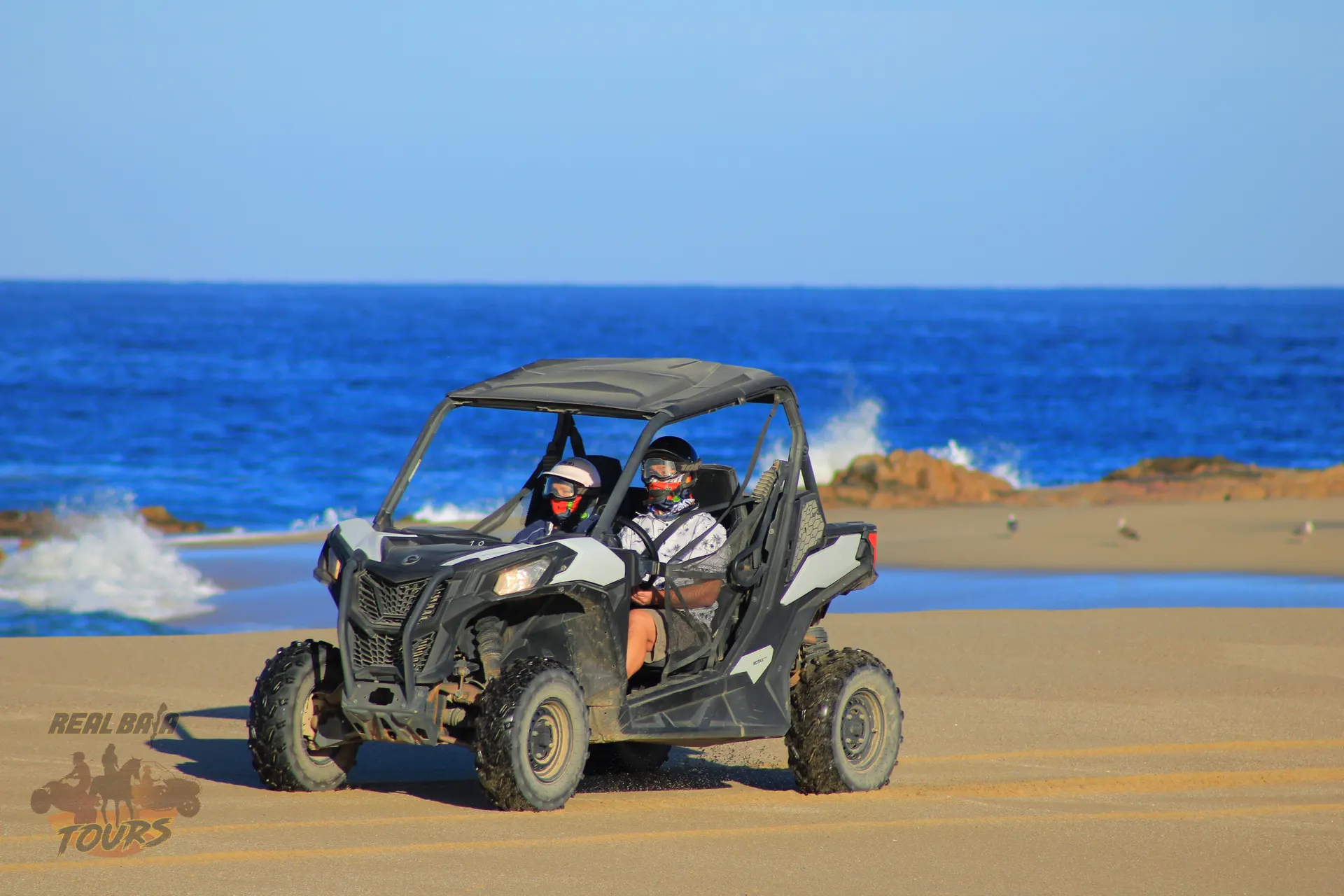 Dos personas manejando a side-by-side UTV on a playa de arena with olas del océano and rocky outcrops in Baja California Sur