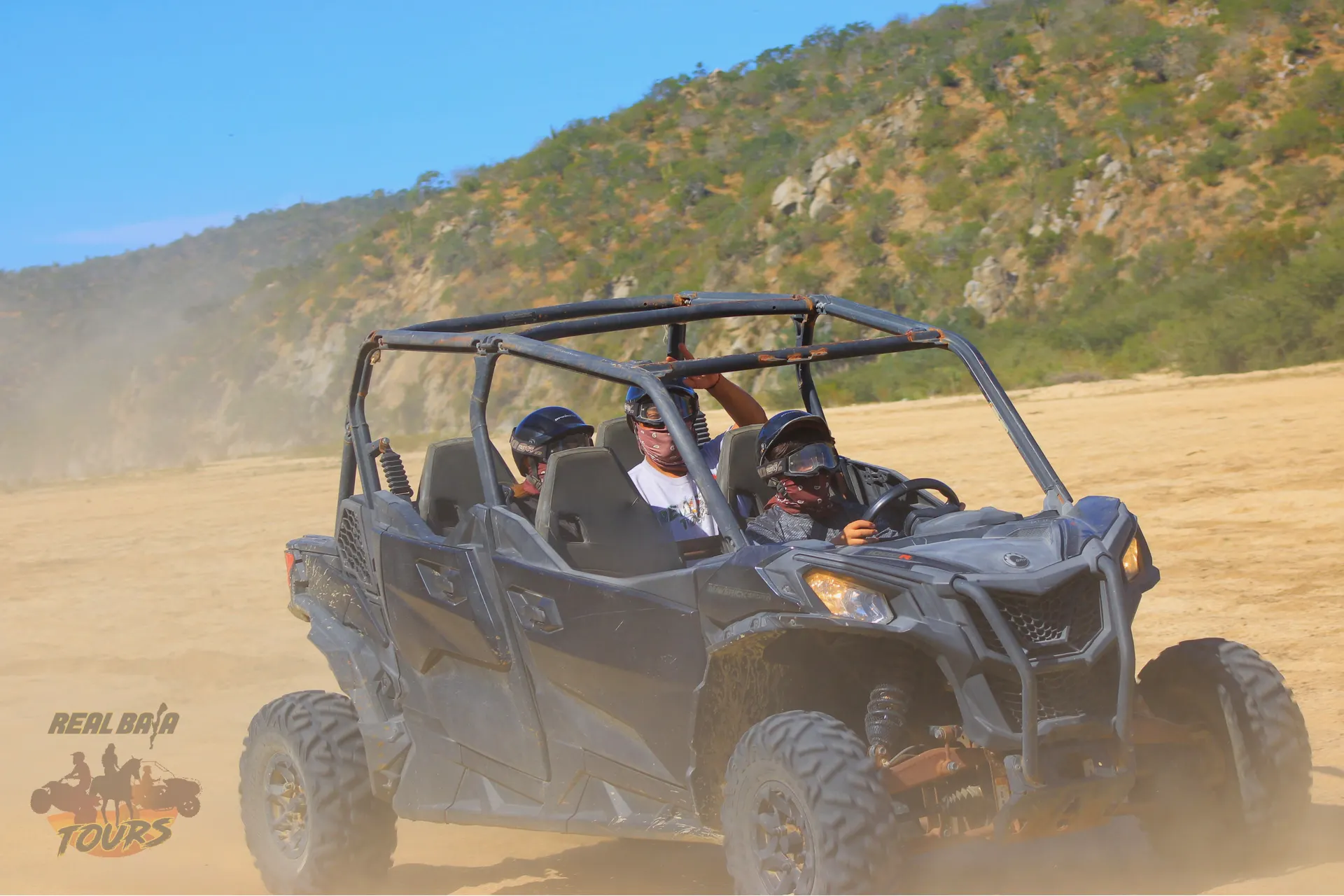 3 tourist manejando in a black open-air UTV across dusty terreno desértico with rocky hills in background near Los Cabos, Baja Cali
