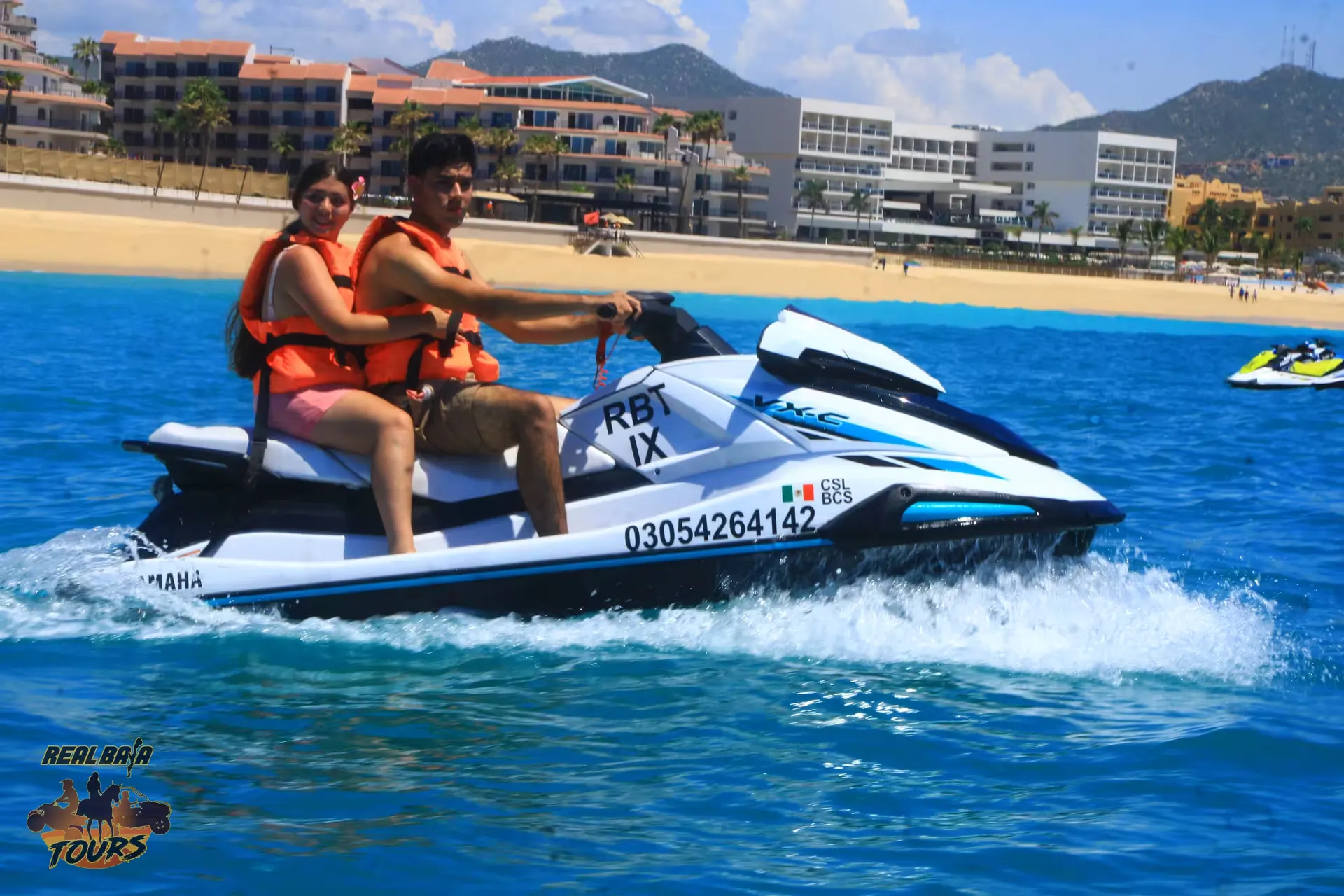 Two people wearing orange life jackets riding a white Yamaha jet ski in turquoise ocean water near a sandy beach with resort buildings and mountains in background
