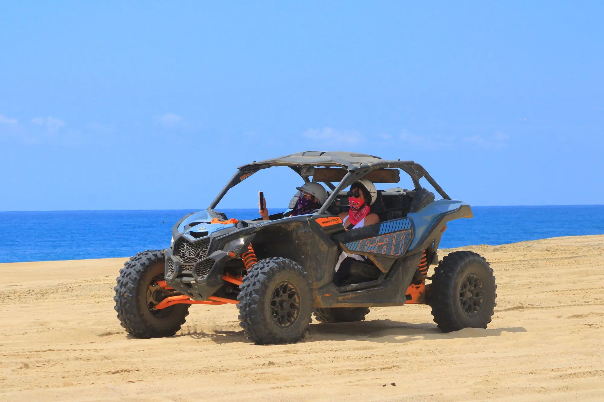 Can-Am Maverick X3 Turbo crossing a coastal sand dune with two women passengers, one taking a photo with her phone, deep blue ocean stretching behind