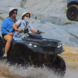 Two riders on an ATV splashing through a shallow water crossing at the base of a granite rock wall, spray kicking up around the tires