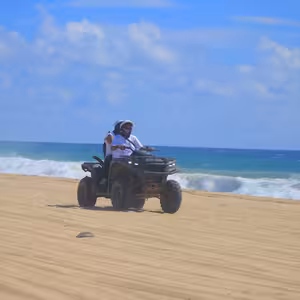 An ATV with two riders cruising along a wide sandy beach as turquoise waves break along the shoreline behind them