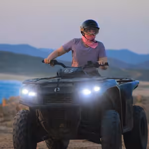 ATV rider with headlights glowing at dusk, coastline and silhouetted mountains stretching behind under a soft blue-gold sky