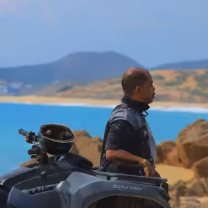 Tour rider standing beside his parked ATV, gazing out over a turquoise bay ringed by desert hills and distant mountains