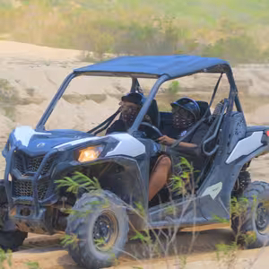 Two riders in a UTV driving through sand dunes with green hills behind