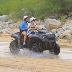 ATV with two riders splashing through a stream lined with boulders
