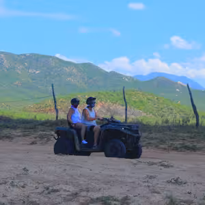 Two riders on an ATV driving through green hills with mountains behind