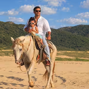 Father and daughter riding a horse together on a sandy trail with mountains in Baja California Sur
