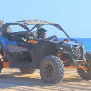Blue and orange Can-Am Maverick X3 Turbo parked on a beach beside dramatic rock formations near Cabo San Lucas, turquoise ocean in the background