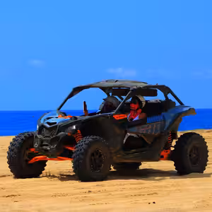Can-Am Maverick X3 Turbo crossing a coastal sand dune with two women passengers, one taking a photo with her phone, deep blue ocean stretching behind