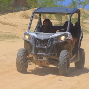 UTV with two riders driving through a sandy dust trail lined with green trees