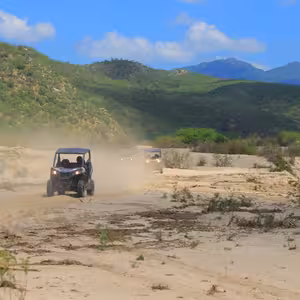 Convoy of UTVs driving through a dusty desert wash with green mountains in the background