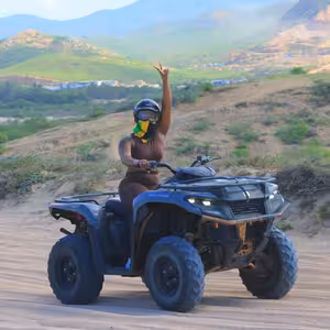 Excited female ATV rider throwing her arm up in celebration on a hillside trail overlooking a green Baja valley and village at golden hour