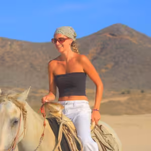 Close-up portrait of a smiling woman on a white horse, wearing a bandana and sunglasses, with dry desert hills in the background
