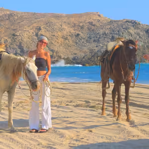Couple standing beside their saddled horses on a sandy beach, smiling at the camera with a rocky headland and turquoise ocean behind them