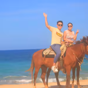 Couple riding double on a horse along the beach, waving and flashing a peace sign under a clear blue sky