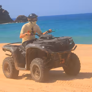 ATV rider on a sandy beach beside a rocky outcrop, with bright turquoise water in the background