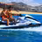 Two people wearing orange life jackets riding a white Yamaha jet ski in turquoise ocean water near a sandy beach with resort buildings and mountains in background